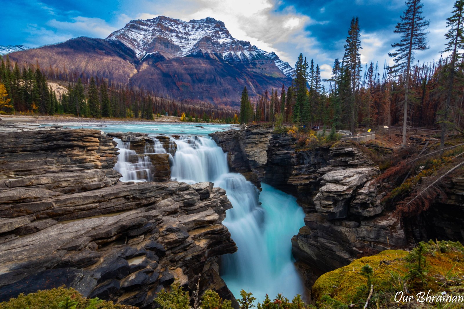 The powerful Athabasca Falls cascades down a rugged, rocky gorge into the turquoise Athabasca River, surrounded by a dense pine forest and towering mountains under a cloudy sky at Jasper National Park, Canada.