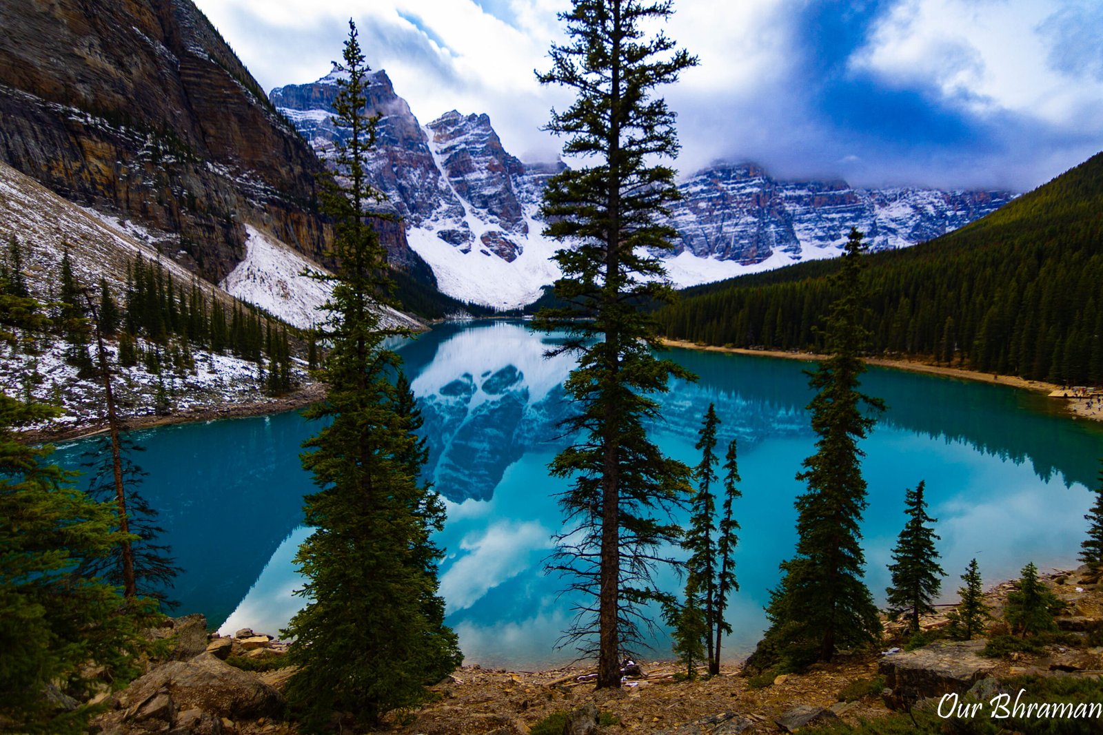 The vibrant turquoise waters of Moraine Lake perfectly reflect the towering, snow-dusted mountains of the Valley of the Ten Peaks, framed by tall evergreen pine trees under a cloudy blue sky at Banff National Park, Canada.