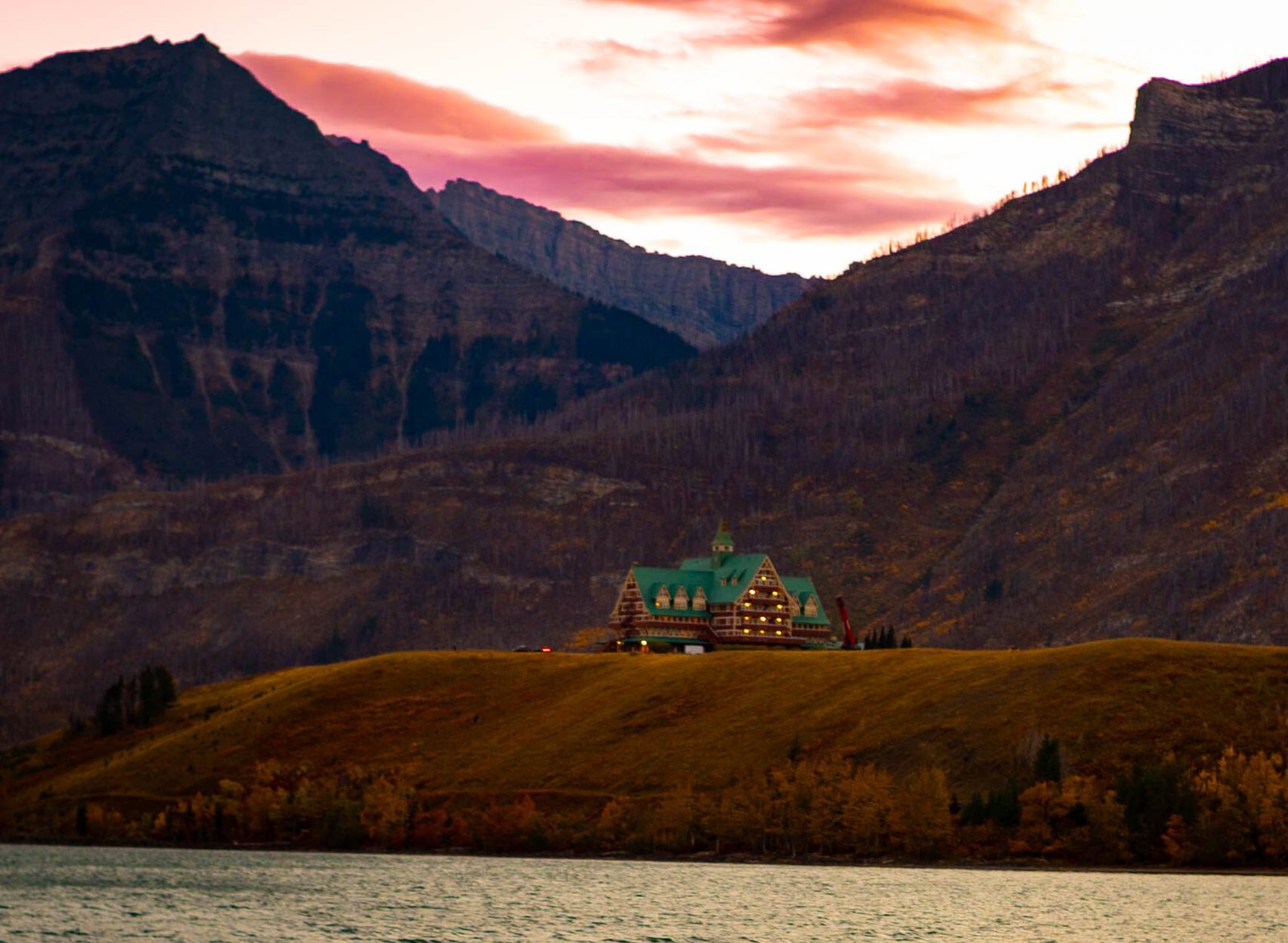 The historic Prince of Wales Hotel stands on a grassy hill overlooking a calm lake, surrounded by towering mountain peaks under a sunset sky at Waterton Lakes National Park, Canada.