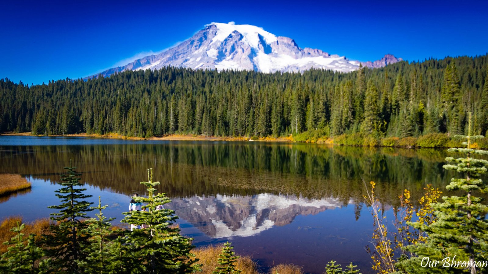 The snow-capped, glacier-covered peak of Mount Rainier reflects perfectly in the calm, dark waters of Reflection Lake