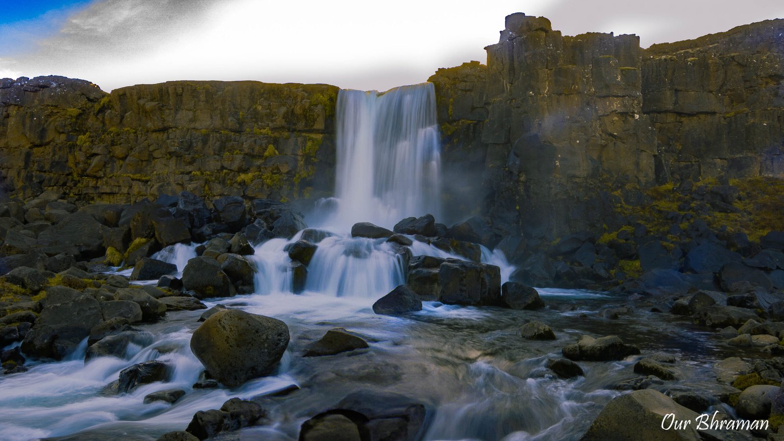 The scenic Öxarárfoss waterfall cascades over a dark, rugged basalt rock cliff into a rocky pool within the Almannagjá gorge at Thingvellir National Park, Iceland.