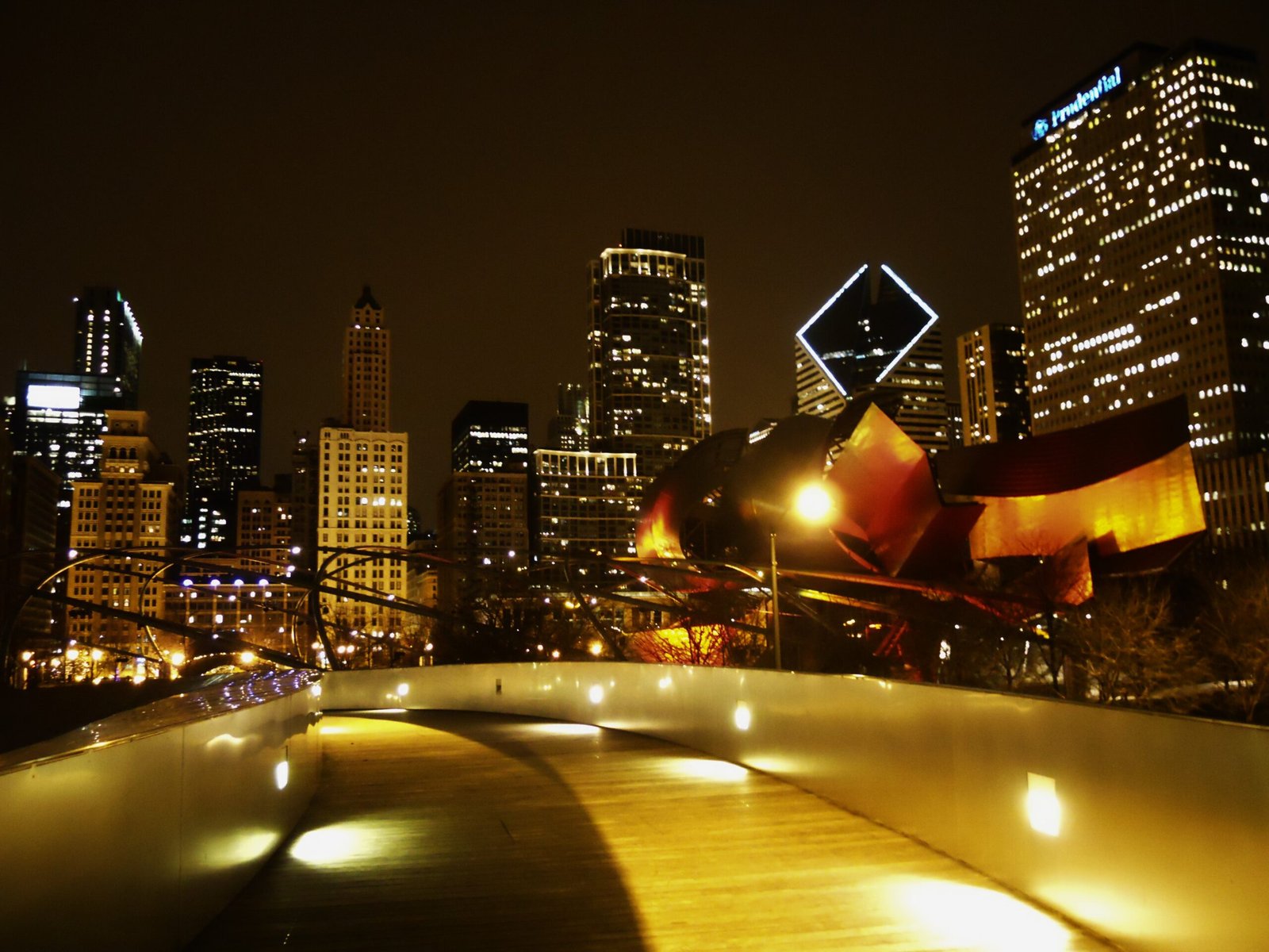Warmly lit curved pedestrian walkways at Millennium Park lead toward the illuminated modern skyscrapers of the downtown Chicago skyline, including the Prudential building, at night.