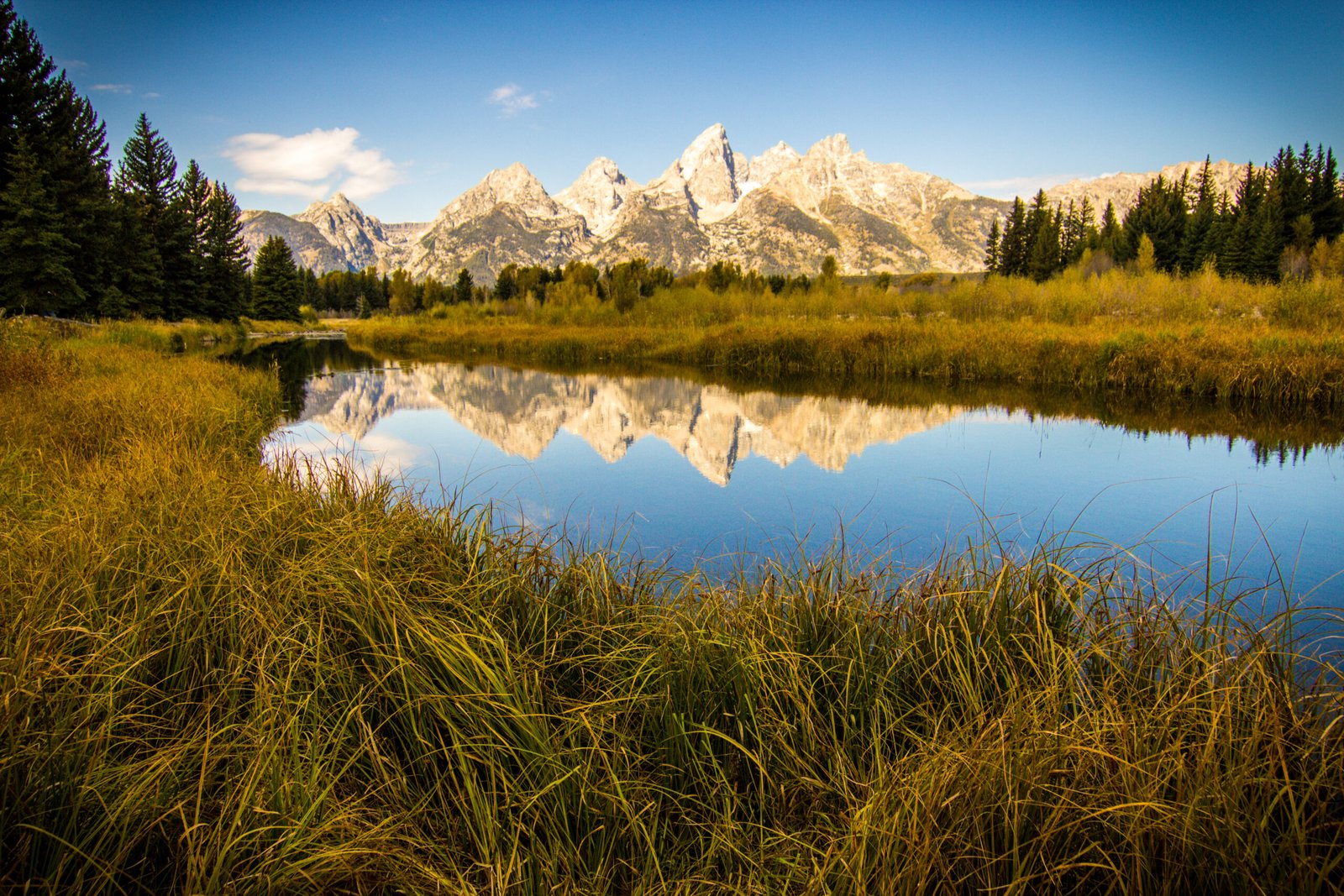The snow-capped mountain peaks of the Grand Teton range are perfectly reflected in the calm, glassy waters of the Snake River