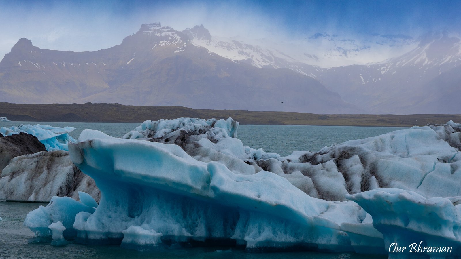 Large, striking blue icebergs float in the calm waters of Jökulsárlón Glacier Lagoon, with the massive Vatnajökull glacier and snow-capped mountains stretching across the horizon in Iceland.