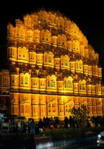 Welcome to Rajasthan The massive, intricately carved red and pink sandstone facade of the Hawa Mahal (Palace of Winds) stands grandly illuminated by warm golden lights at night in Jaipur, India.